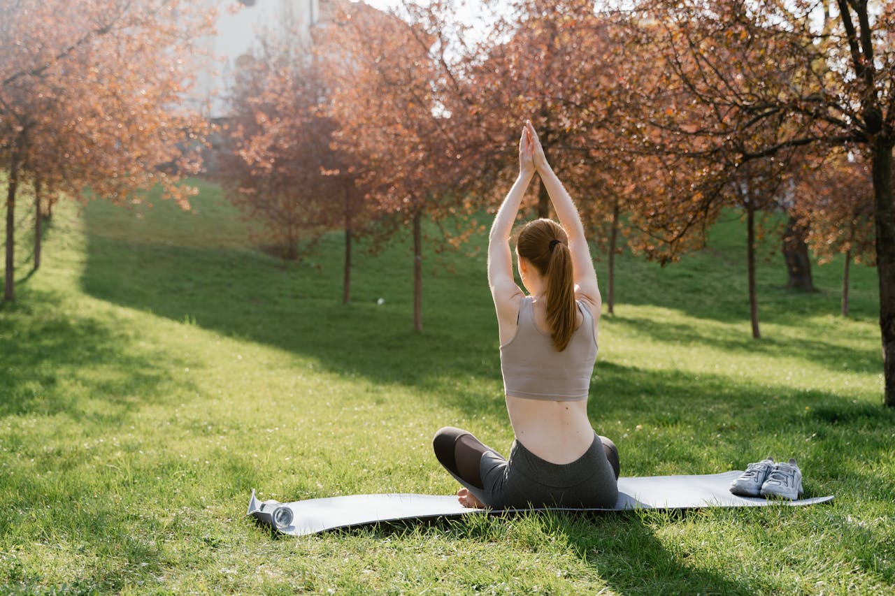 A woman practicing yoga on a mat outdoors in a sunlit park during autumn.